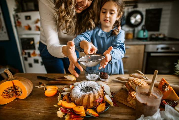 Mother and daughter in a home kitchen sprinkling powdered sugar over a bundt cake. Fall themed decor and pumpkins around the table